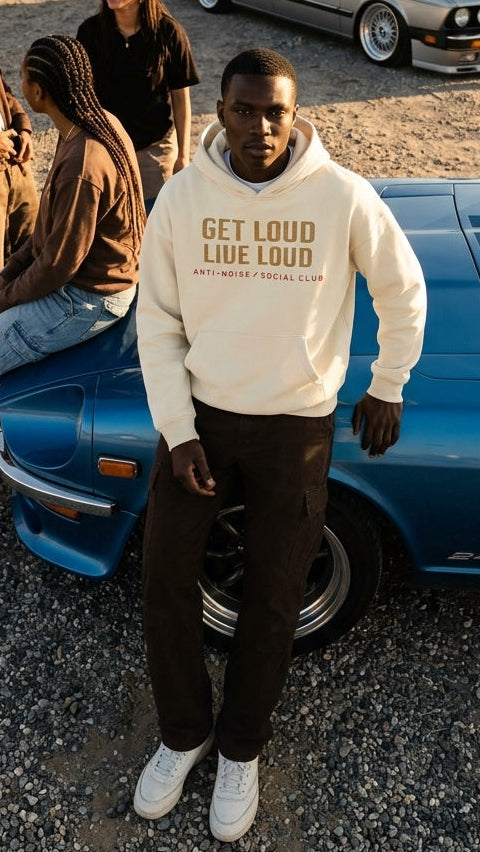 A man wearing a cream hoodie with 'GET LOUD LIVE LOUD' printed on the front stands in front of a blue car in a desert setting, with several people and a silver car in the background.
